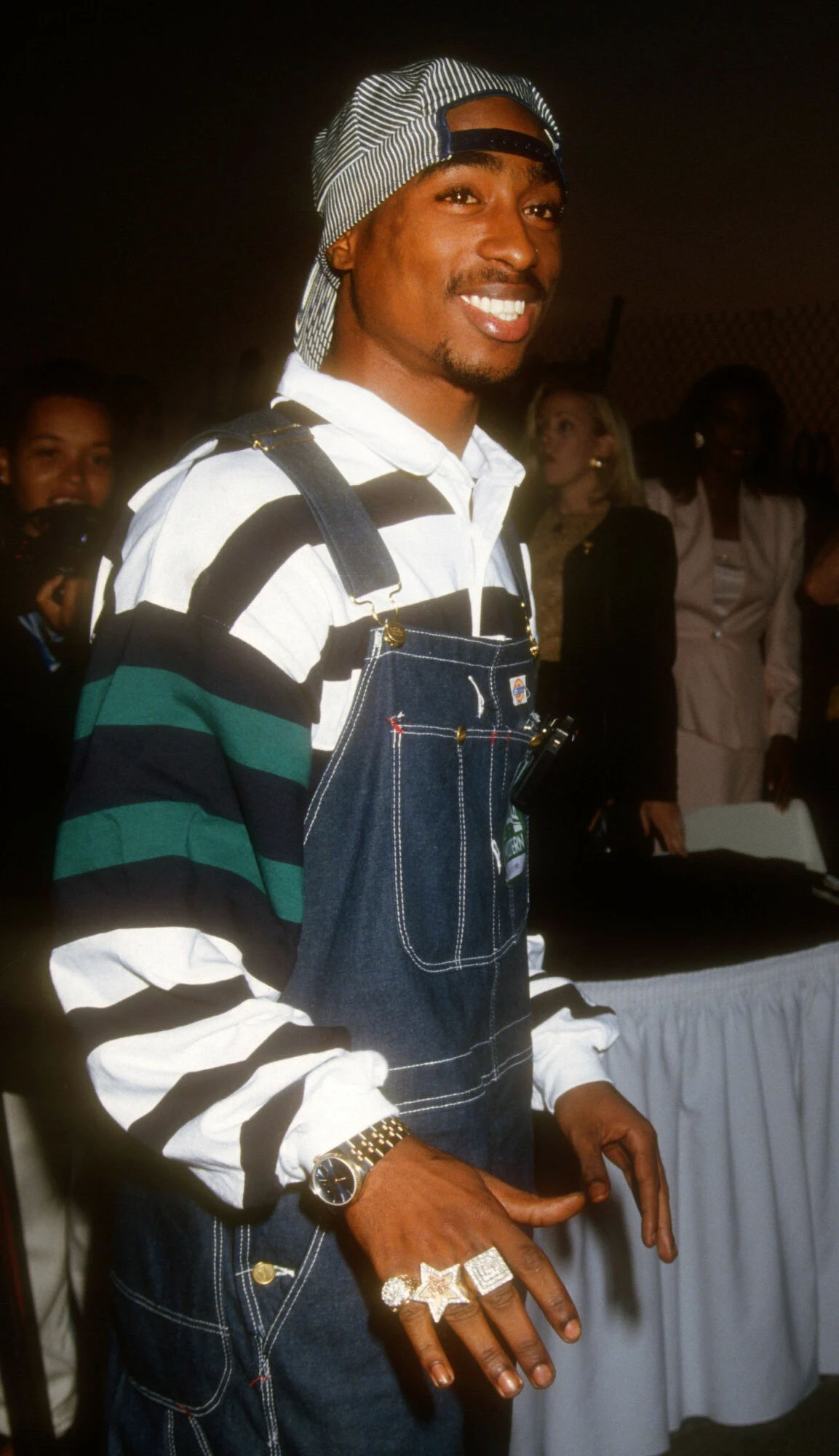 Tupac Shakur Mężczyzna w luźnych ogrodniczkach dżinsowych i pasiastym swetrze z szerokim uśmiechem, ozdobiony pokaźnymi złotymi pierścionkami oraz zegarkiem, stoi na tle grupki elegancko ubranych osób.