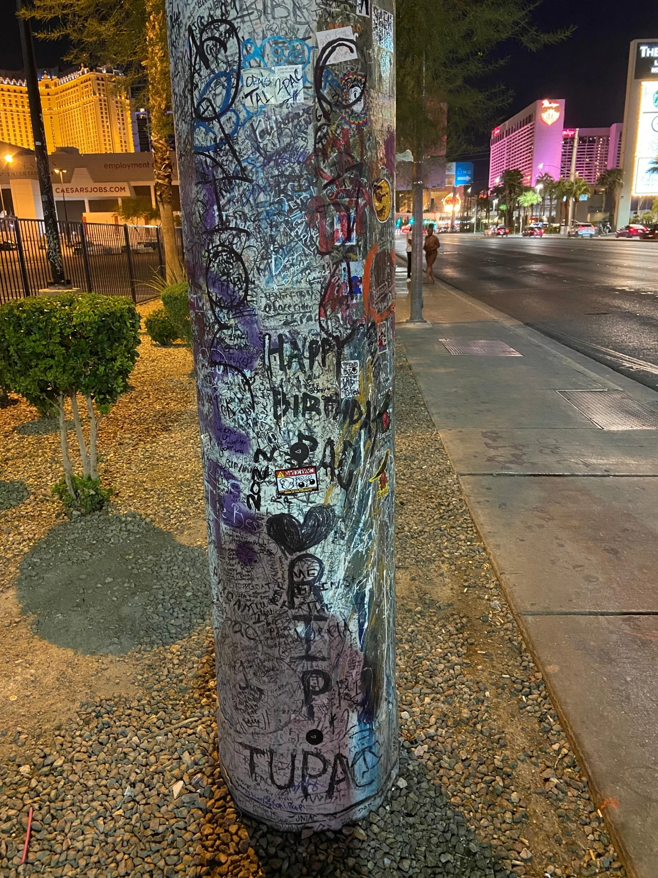 Słup na skrzyżowaniu Flamingo/Koval w Las Vegas, na którym fani Tupaca składają podpisy i wyrazy sympatii wobec muzyka Betonowy słup pokryty różnorodnymi napisami i rysunkami graffiti, wśród których wyróżnia się czarny napis 'R.I.P. TUPAC' oraz kilka serc i podpisów upamiętniających TUPAC-a, znajdujący się przy miejskim chodniku w oświetlonej części ulicy nocą.