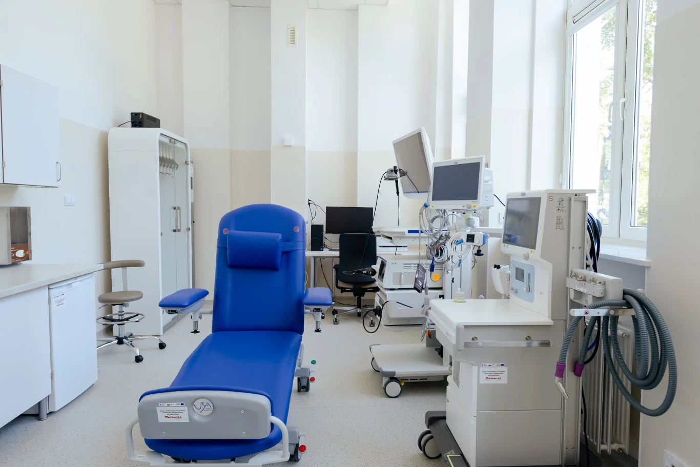 Niebieski fotel medyczny ustawiony centralnie w jasnym, sterylnym pomieszczeniu, otoczony specjalistyczną aparaturą medyczną, monitorem oraz urządzeniami diagnostycznymi przy dużym oknie.