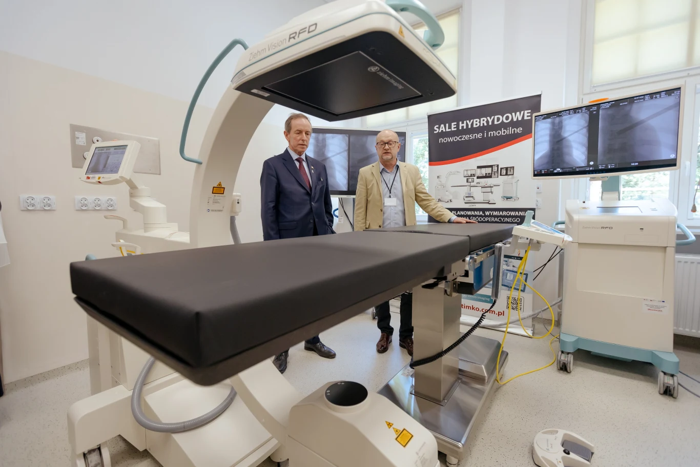 Dwóch mężczyzn w garniturach stoi obok nowoczesnego stołu zabiegowego w sali hybrydowej wyposażonej w zaawansowany sprzęt medyczny, ekrany oraz aparaturę obrazującą, na tle banera informacyjnego.