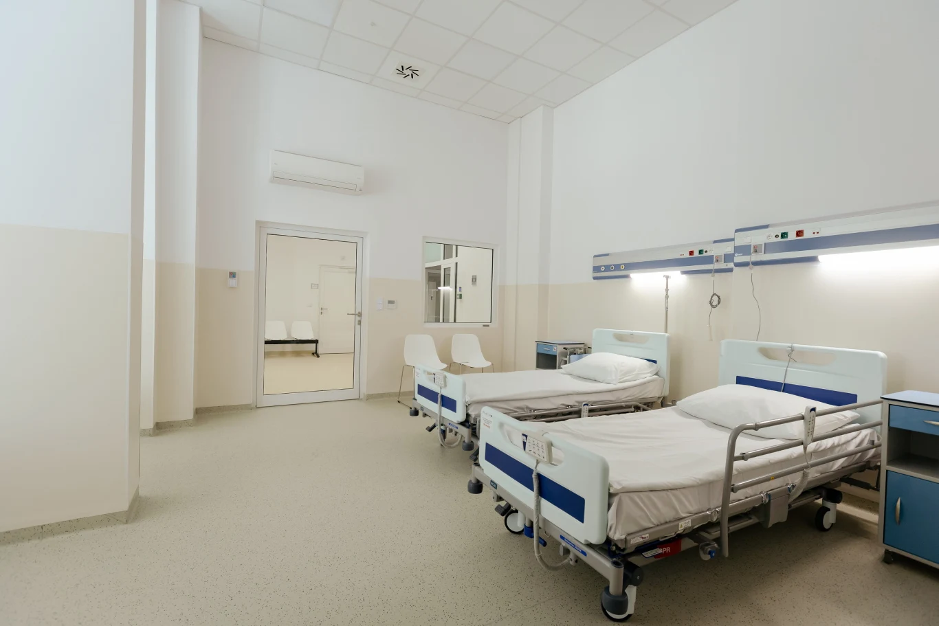 Dwa puste łóżka szpitalne z białą pościelą, stojące obok siebie przy ścianie z panelami do monitorowania stanu zdrowia, jasne wnętrze z wysokim sufitem i dużym oknem w drzwiach.