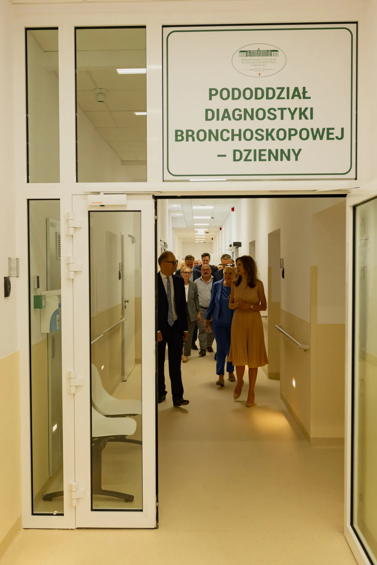 Grupa osób przechodzi przez jasny korytarz szpitalny w kierunku wyjścia z pomieszczenia oznaczonego jako pododdział diagnostyki bronchoskopowej.