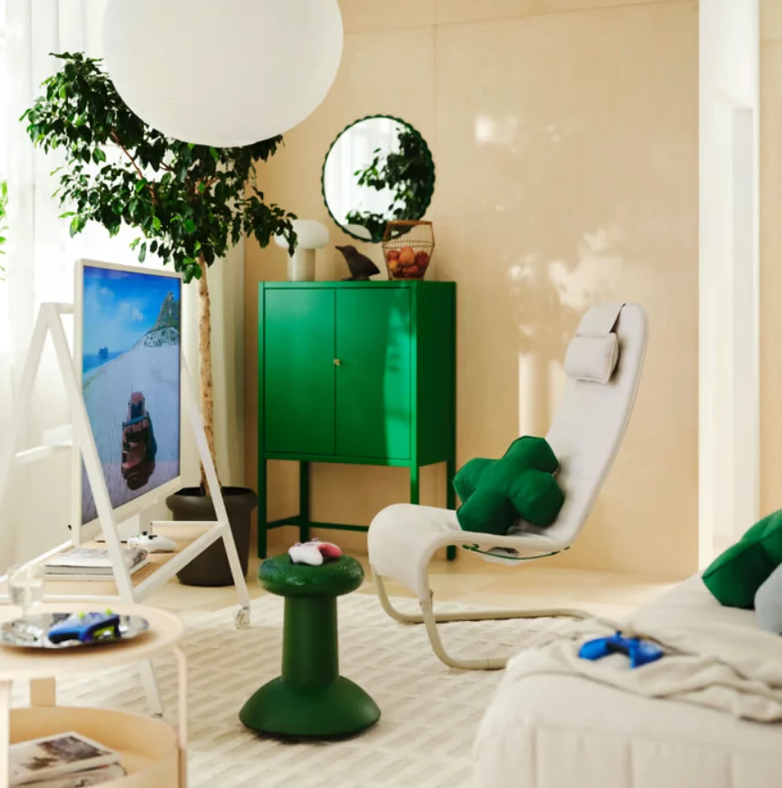 IKEA x Xbox Jasny salon urządzony w nowoczesnym stylu z białym fotelem relaksacyjnym i zielonymi akcentami, telewizorem ustawionym na sztaludze oraz zieloną szafką na tle beżowych ścian, uzupełniony dużą rośliną i lustrem nad meblem.