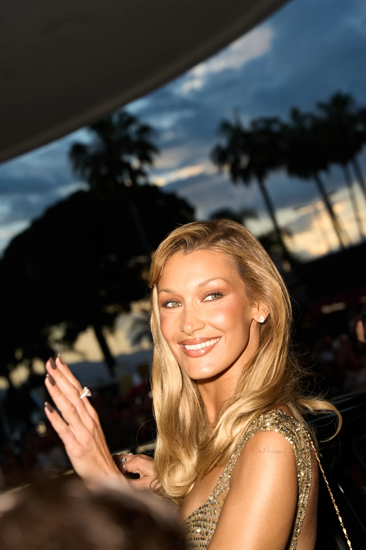 Bella Hadid i jej "honey blonde". Koloryzacja nadaje cerze blasku Kobieta z długimi blond włosami, ubrana w błyszczącą suknię, uśmiecha się i macha ręką, w tle sylwetki palm i wieczorne niebo.