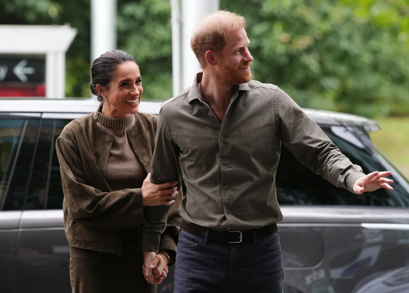 Meghan i Harry wylądowali już na lotnisku w Birmingham Uśmiechnięta para trzymająca się za ręce, kobieta ubrana w brązową kurtkę, mężczyzna w oliwkowej koszuli, stoją obok ciemnego samochodu, w tle zielone drzewa.