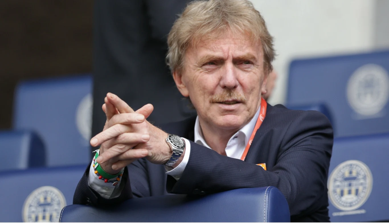 Zbigniew Boniek Mężczyzna w garniturze i białej koszuli siedzi na stadionowej trybunie, opierając ręce o przednie oparcie, z poważnym wyrazem twarzy skierowanym w stronę boiska.