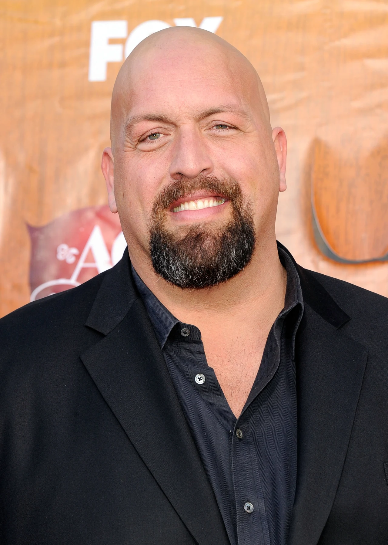 Wrestler Paul Wight Łysy mężczyzna z brodą ubraną w czarną koszulę i czarną marynarkę, uśmiechnięty, stoi na tle brązowej ścianki z logotypami.