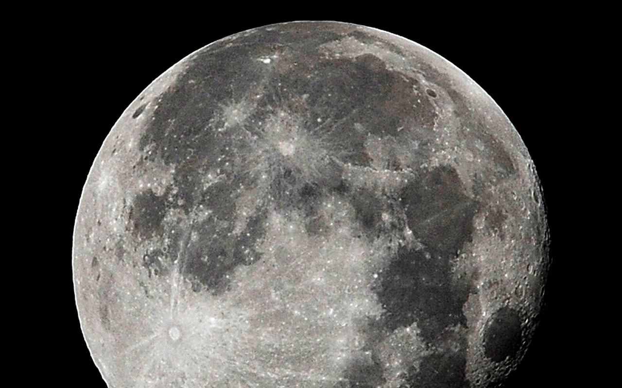 Księżyc Zbliżenie jasnej powierzchni Księżyca z wyraźnie widocznymi kraterami i ciemnymi plamami, na tle czarnego nieba.