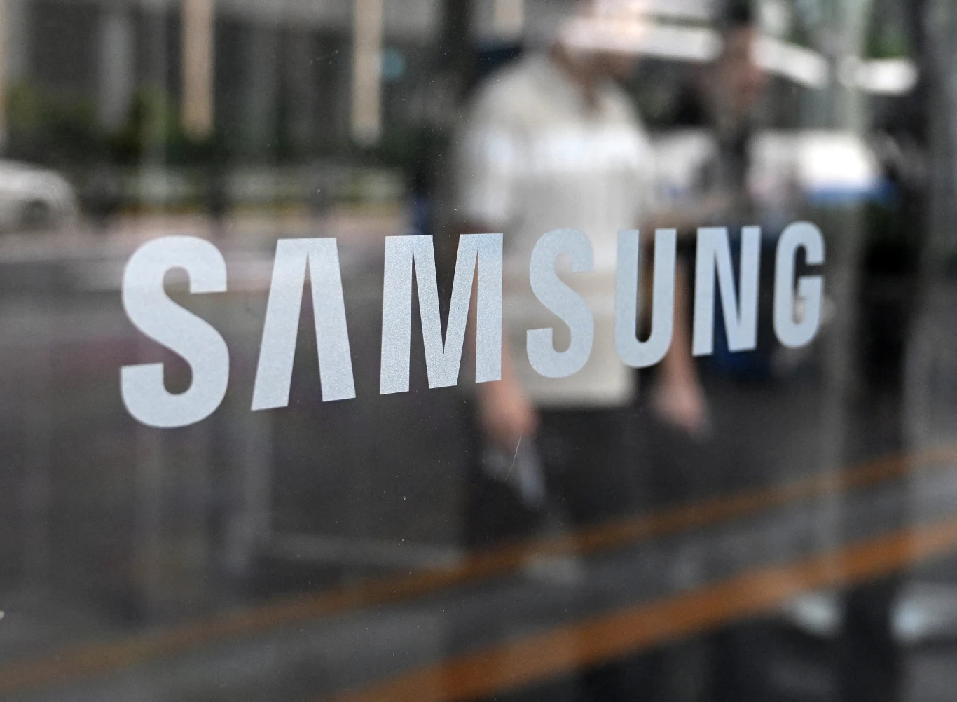 Samsung Logo Samsung na szklanych drzwiach, w tle sylwetki ludzi i fragment ulicy za szybą.