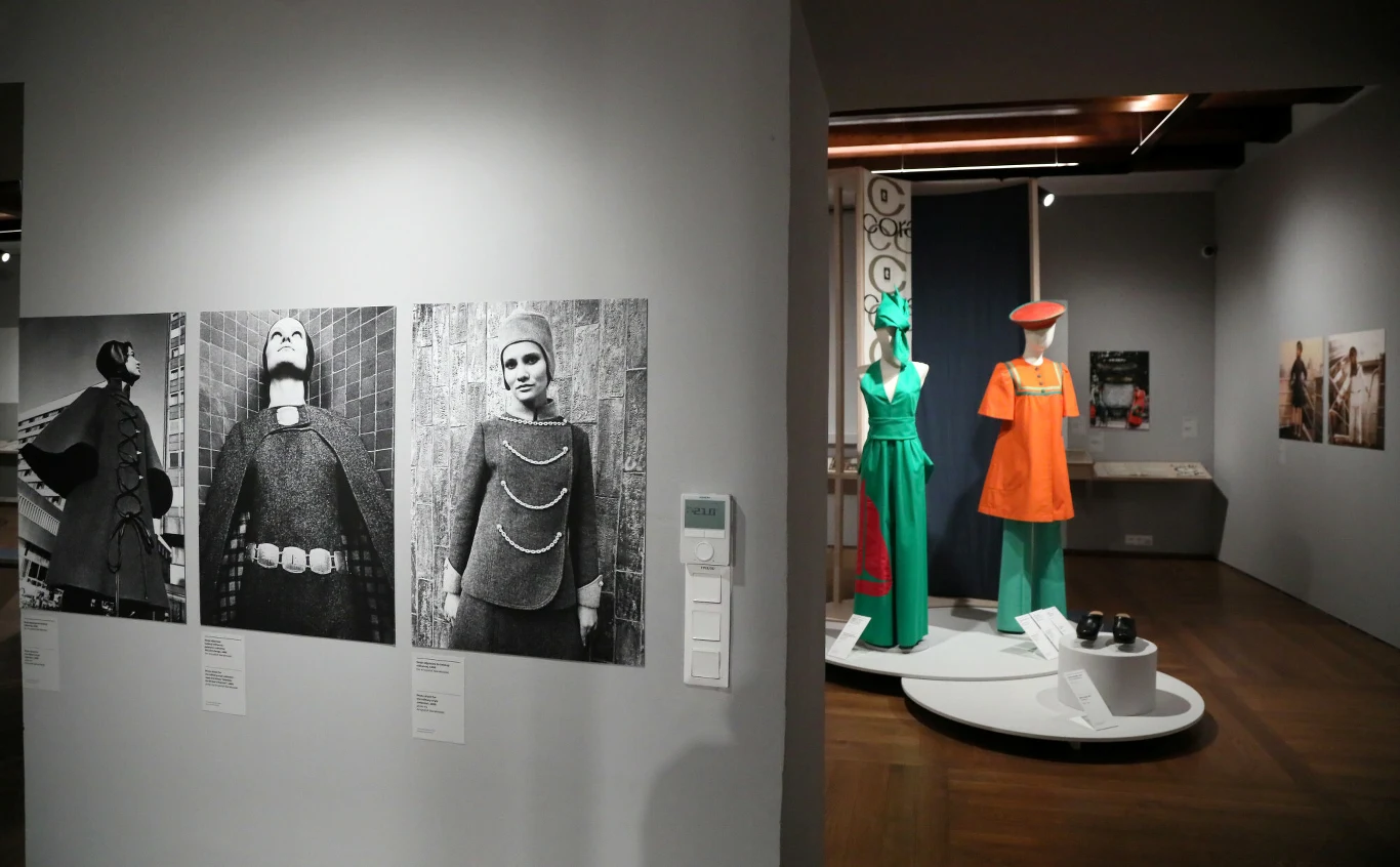 Wystawa "Grazyna Hase. Zawsze w modzie" w Muzeum Warszawy. Czarno-białe fotografie przedstawiające osoby w awangardowych stylizacjach oraz manekiny ubrane w kolorowe stroje retro, ustawione na białym podwyższeniu w muzealnej przestrzeni.