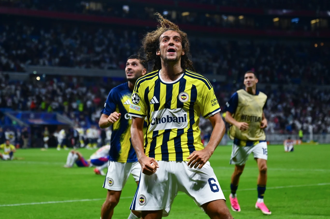 Matteo Guendouzi Piłkarz Fenerbahce w żółto-granatowej koszulce z numerem 6 wyraża radość po zdobyciu gola, z zaciśniętymi pięściami i rozwianymi włosami, otoczony przez współprowadzących go kolegów z drużyny na tle rozemocjonowanych kibiców na stadionie.