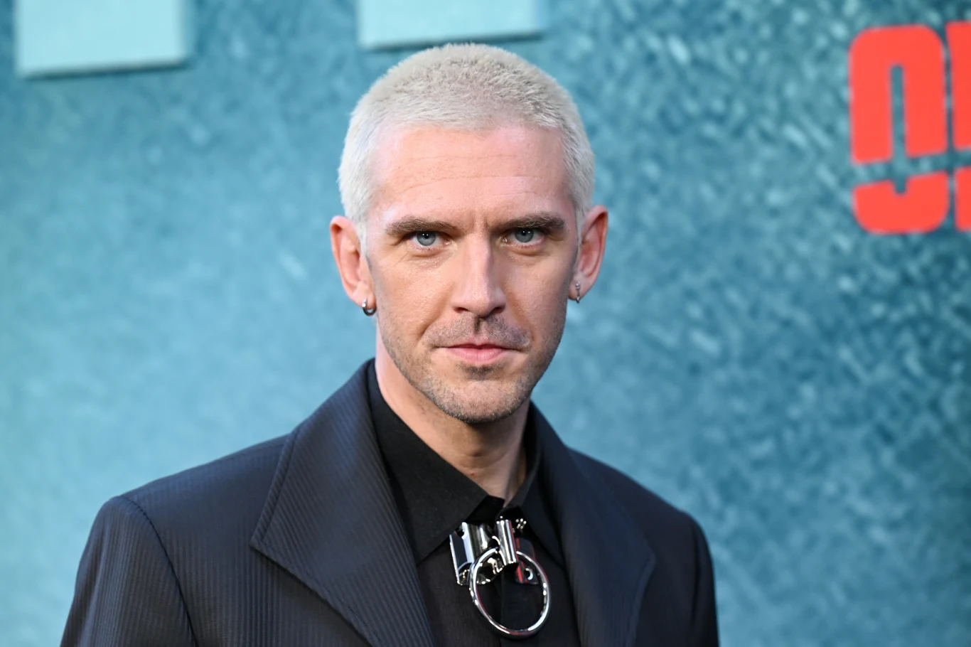 Dan Stevens w 2026 roku Dan Stevens w 2026 roku w czarnym garniturze, z metalową biżuterią na tle niebieskiej ściany z czerwonym napisem.