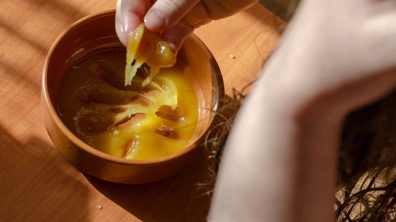 Miód może przydać się poza kuchnią. To jeden z naturalnych składników stosowanych w domowej pielęgnacji włosów. Ręka zanurzająca się w misce z miodem, stojącej na drewnianym stole.