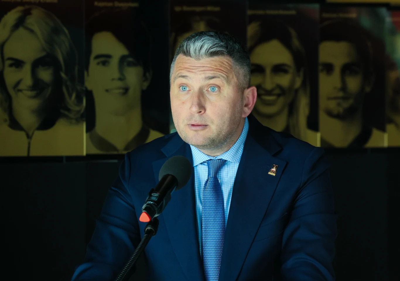 Mężczyzna w garniturze i niebieskim krawacie przemawia do mikrofonu na tle dużych czarno-białych portretów osób.