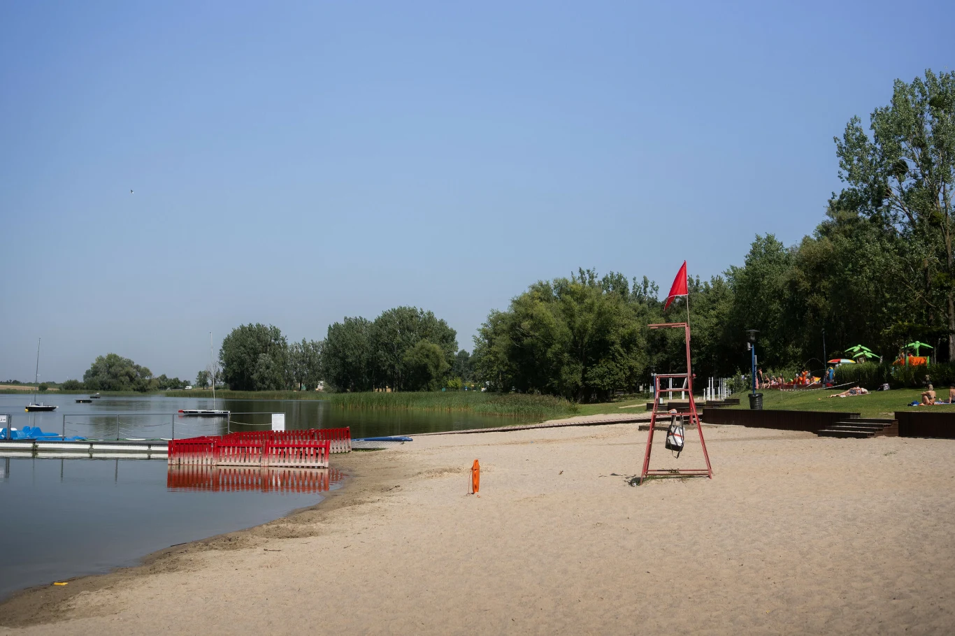 Poziom wody w jeziorze spadł o ok. 1,5 metra Piaszczysta plaża z wieżą ratownika i czerwoną flagą przy sztucznie wydzielonym kąpielisku, otoczona zielonymi drzewami i trawnikiem.