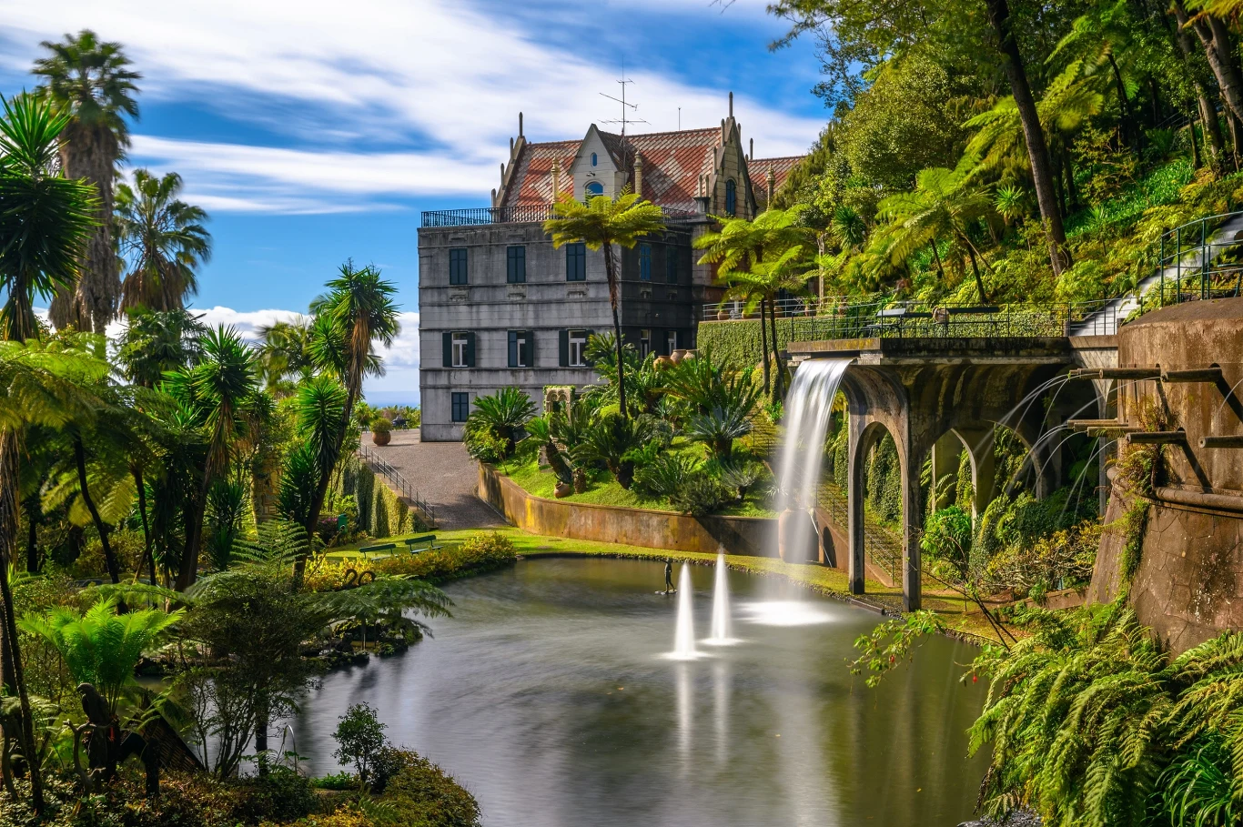 Fontanna w ogrodzie Monte Palace w Funchal na Maderze Historyczny budynek o czerwonym dachu otoczony bujną, tropikalną roślinnością, nad malowniczym stawem z wodospadem i fontannami.