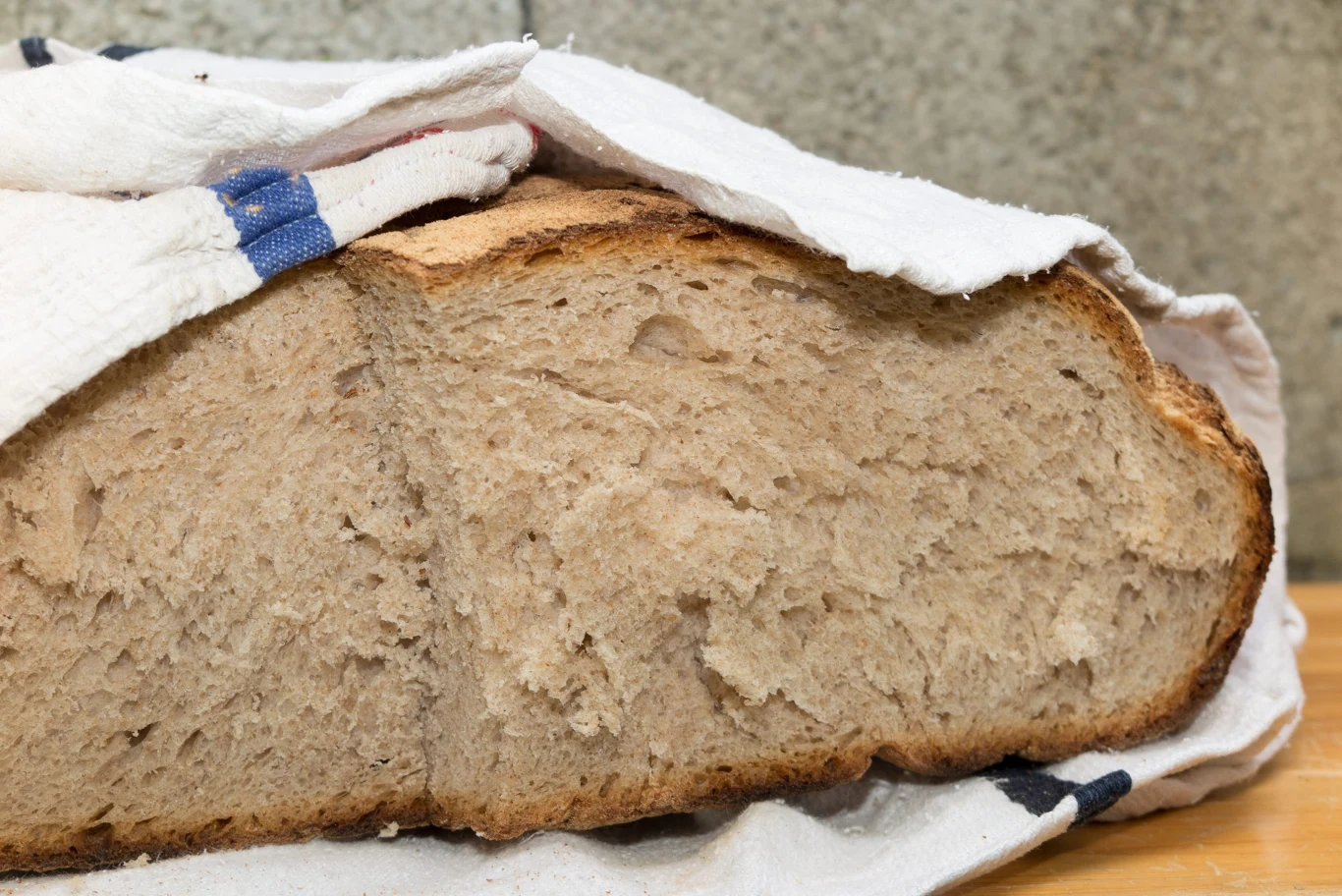 Kromka domowego chleba przykryta białą ściereczką z niebieskimi paskami, ułożona na drewnianej powierzchni.