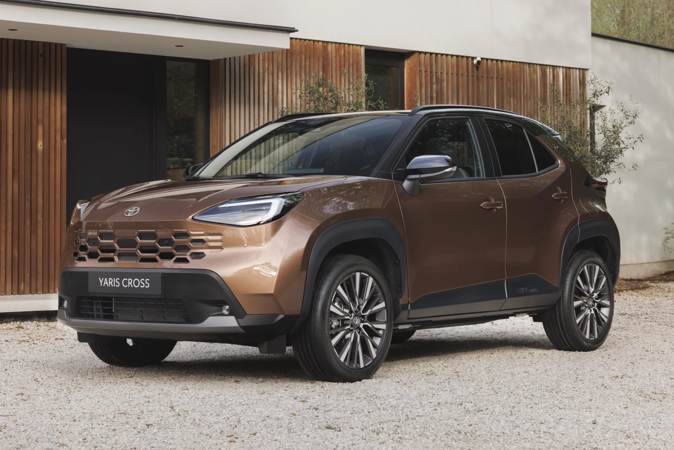 Bestseller Toyoty tańszy. Pięć wersji dostało specjalną cenę Toyota Yaris Cross w kolorze brązowym na podjeździe przed nowoczesnym budynkiem z białymi i drewnianymi fasadami.