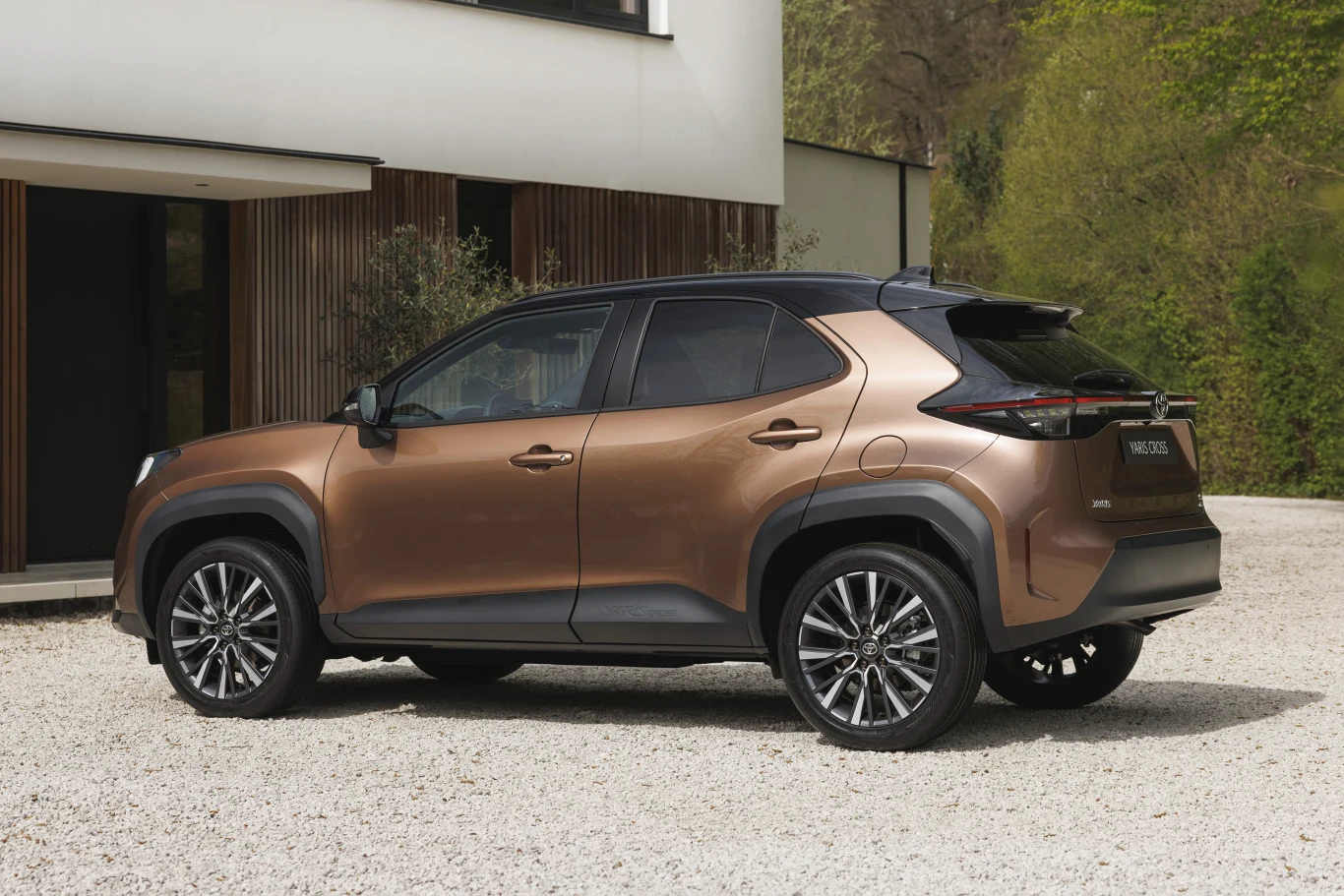 Pod maską Yaris Cross pozostała hybryda oparta na silniku 1.5 Hybrid Dynamic Force. Brązowy, nowoczesny crossover z czarnym dachem i masywnymi nadkolami zaparkowany na żwirowym podjeździe obok minimalistycznego, jasnego budynku.