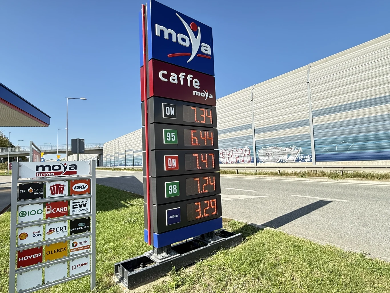 Ceny paliw na stacji Moya 27 sierpnia Ceny paliw na stacji Moya, tablica z aktualnymi kwotami za benzynę, olej napędowy oraz LPG przy ruchliwej drodze, w tle nowoczesne ekrany akustyczne.