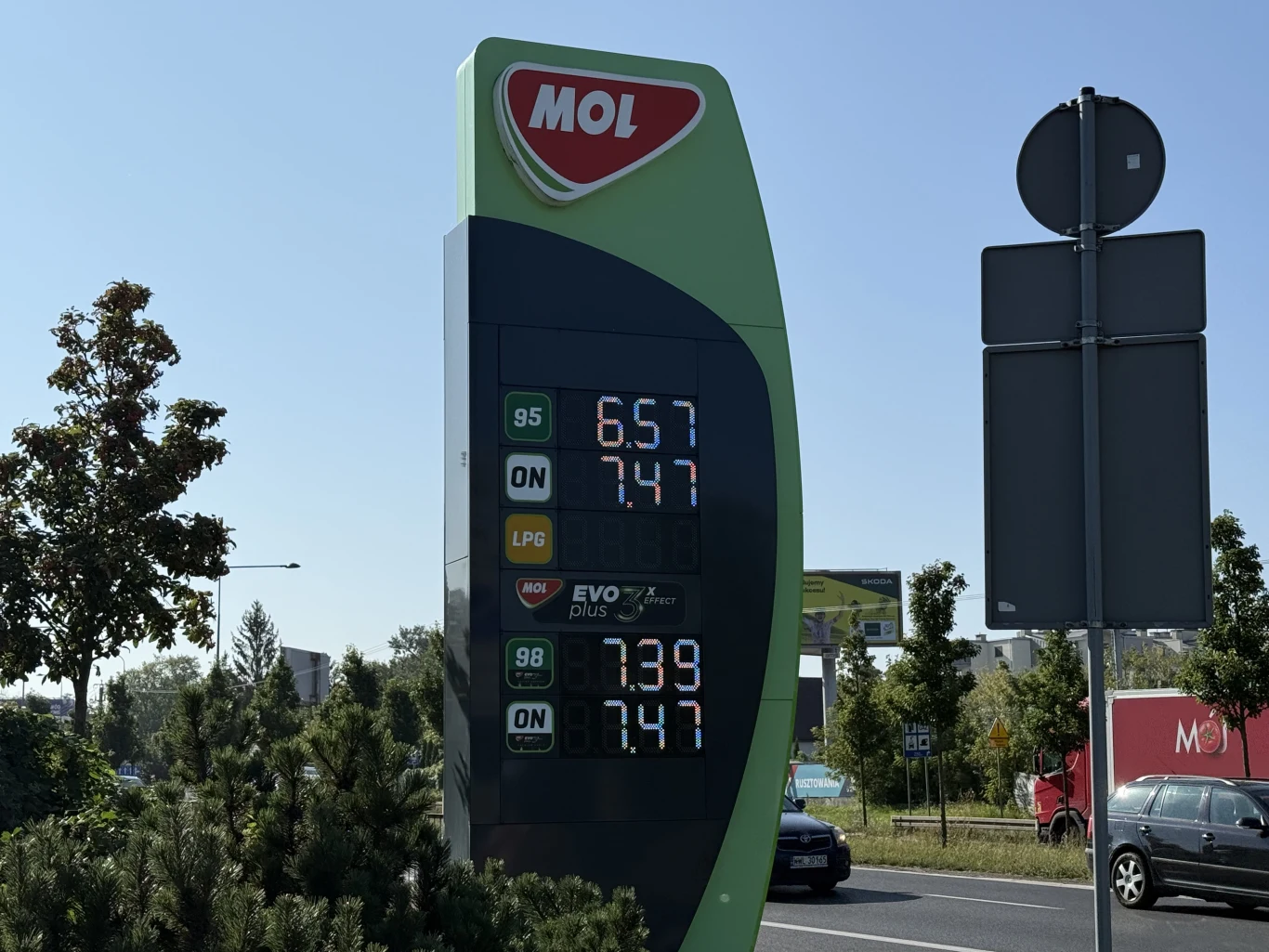 Ceny paliw na stacji MOL 27 sierpnia Tablica cenowa stacji paliw MOL z wyświetlonymi aktualnymi cenami paliw 95, 98, ON i LPG, otoczona zielenią przy ruchliwej ulicy.
