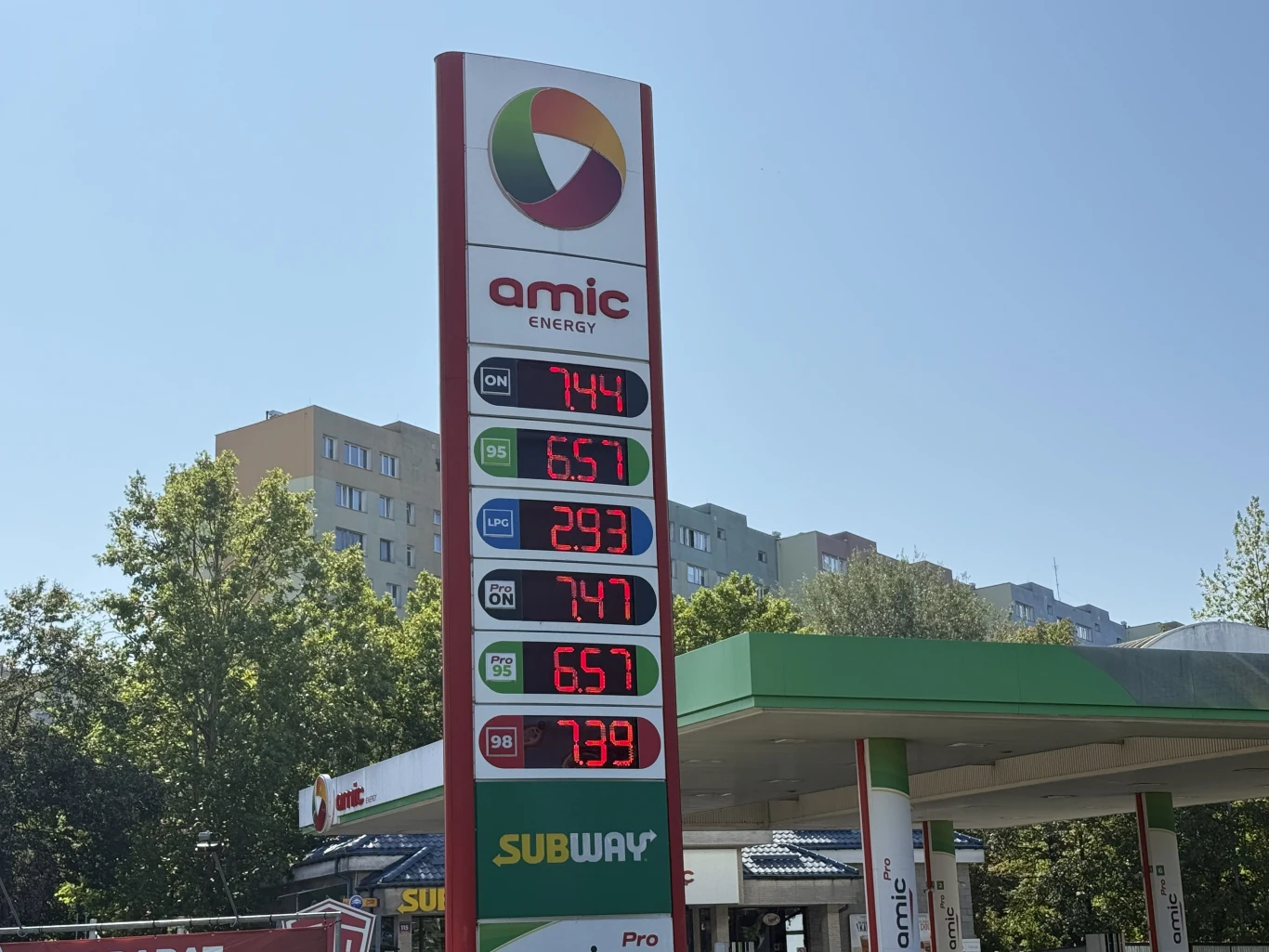 Ceny paliw na stacji Amic 27 sierpnia Słupek cenowy stacji paliw Amic Energy z wyraźnymi cenami różnych rodzajów paliw na tle zielonego daszka stacji i bloków mieszkalnych.