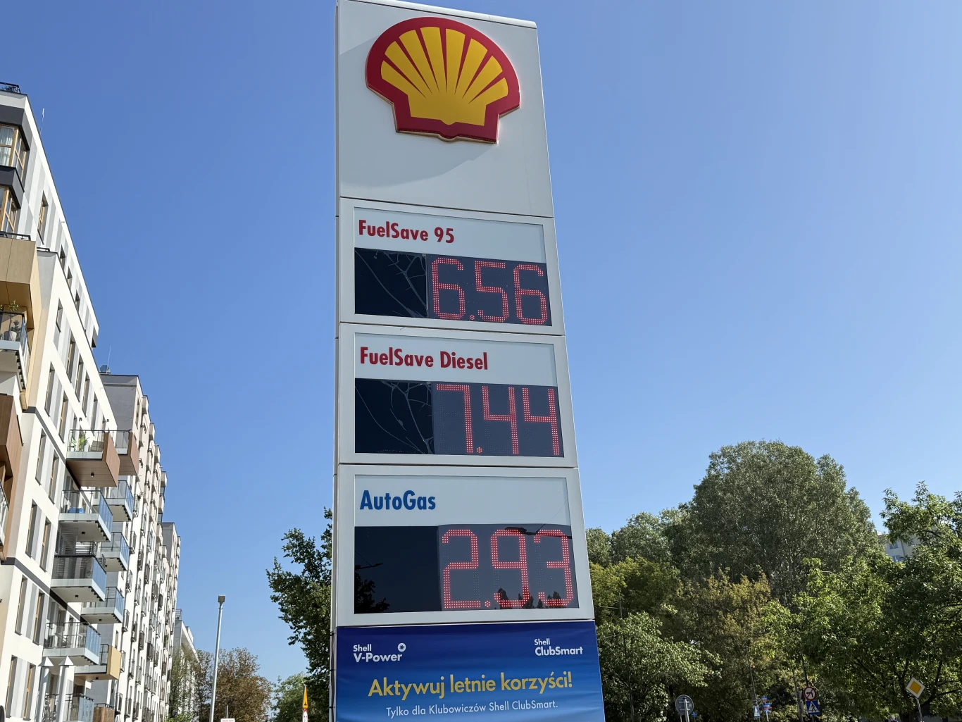 Ceny paliw na Shellu 27 sierpnia Tablica cenowa stacji paliw Shell z wyraźnie widocznymi aktualnymi cenami FuelSave 95, FuelSave Diesel oraz AutoGas na tle nieba i drzew.