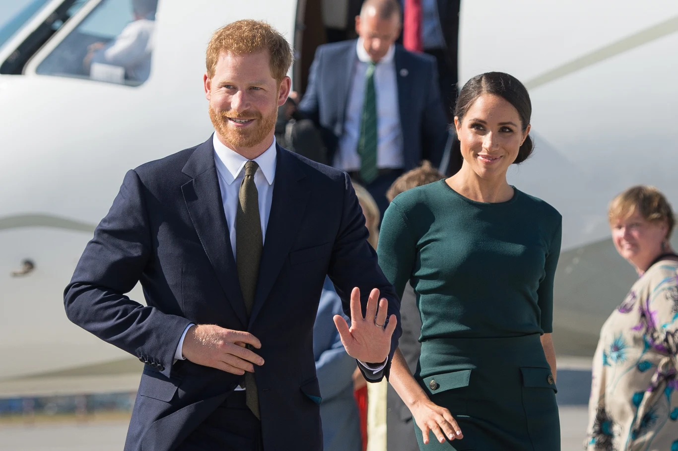 Z czym od początku muszą mierzyć się Sussexowie? Para ubrana w eleganckie stroje wychodzi z samolotu pasażerskiego, uśmiechając się i pozdrawiając zebranych wokół ludzi.