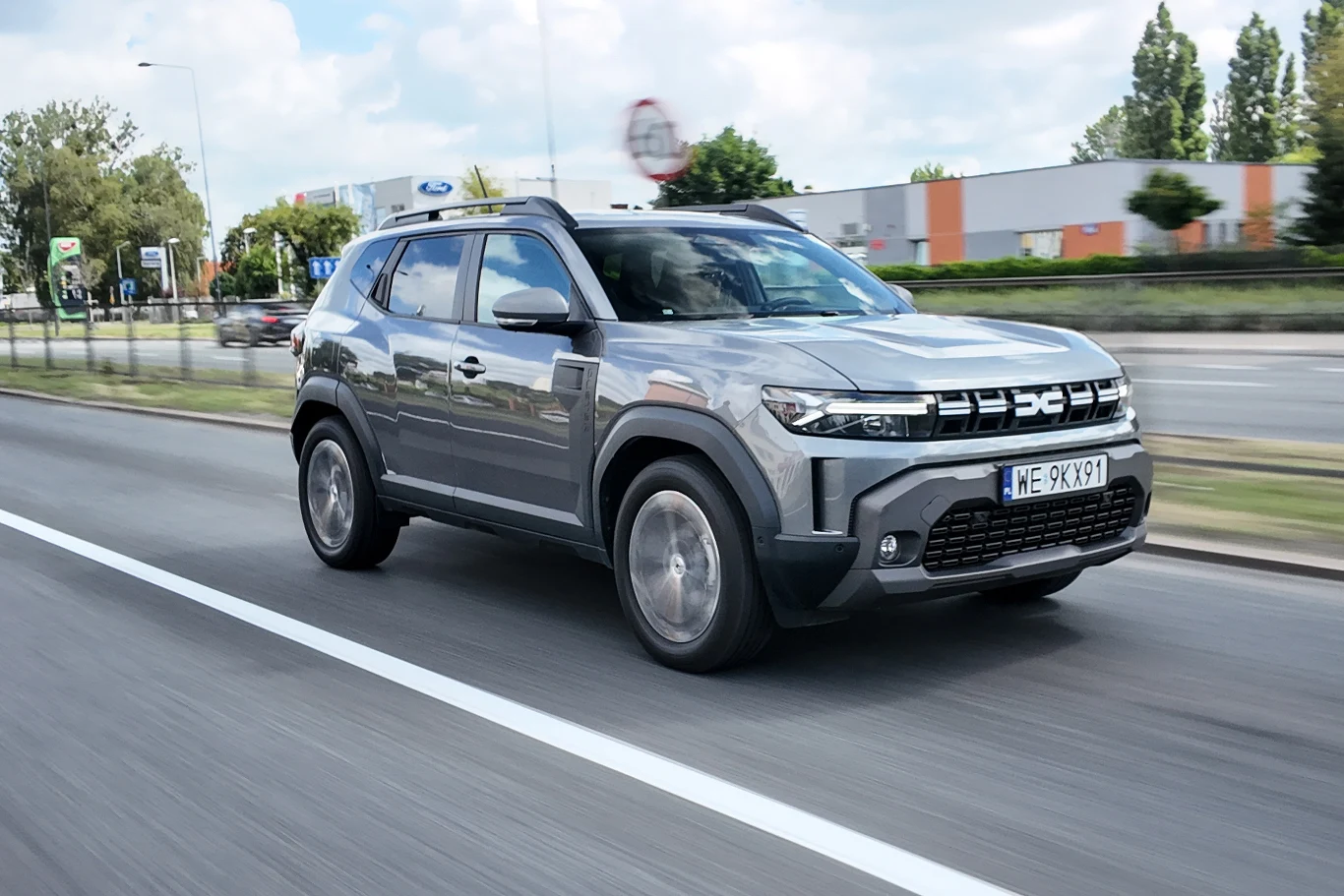 Szary SUV nowej generacji jedzie lewym pasem miejskiej ulicy, wyróżniając się charakterystycznym, nowoczesnym grillem i agresywnymi światłami LED.