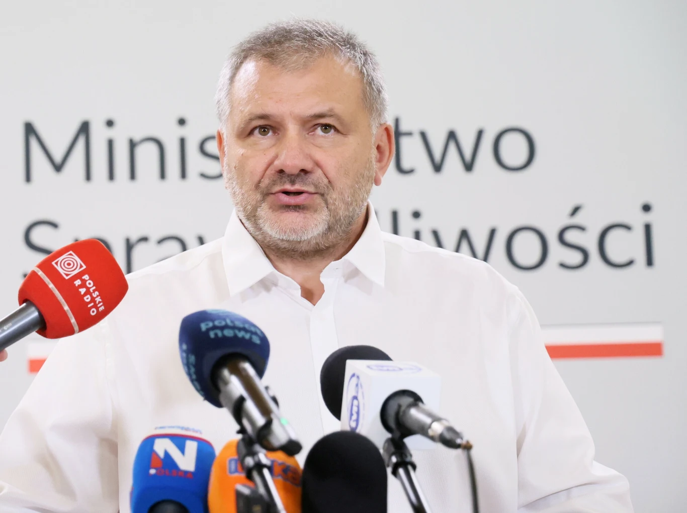 Waldemar Żurek Mężczyzna w białej koszuli przemawia do mediów przy kilku mikrofonach ustawionych na tle napisu Ministerstwo Sprawiedliwości.