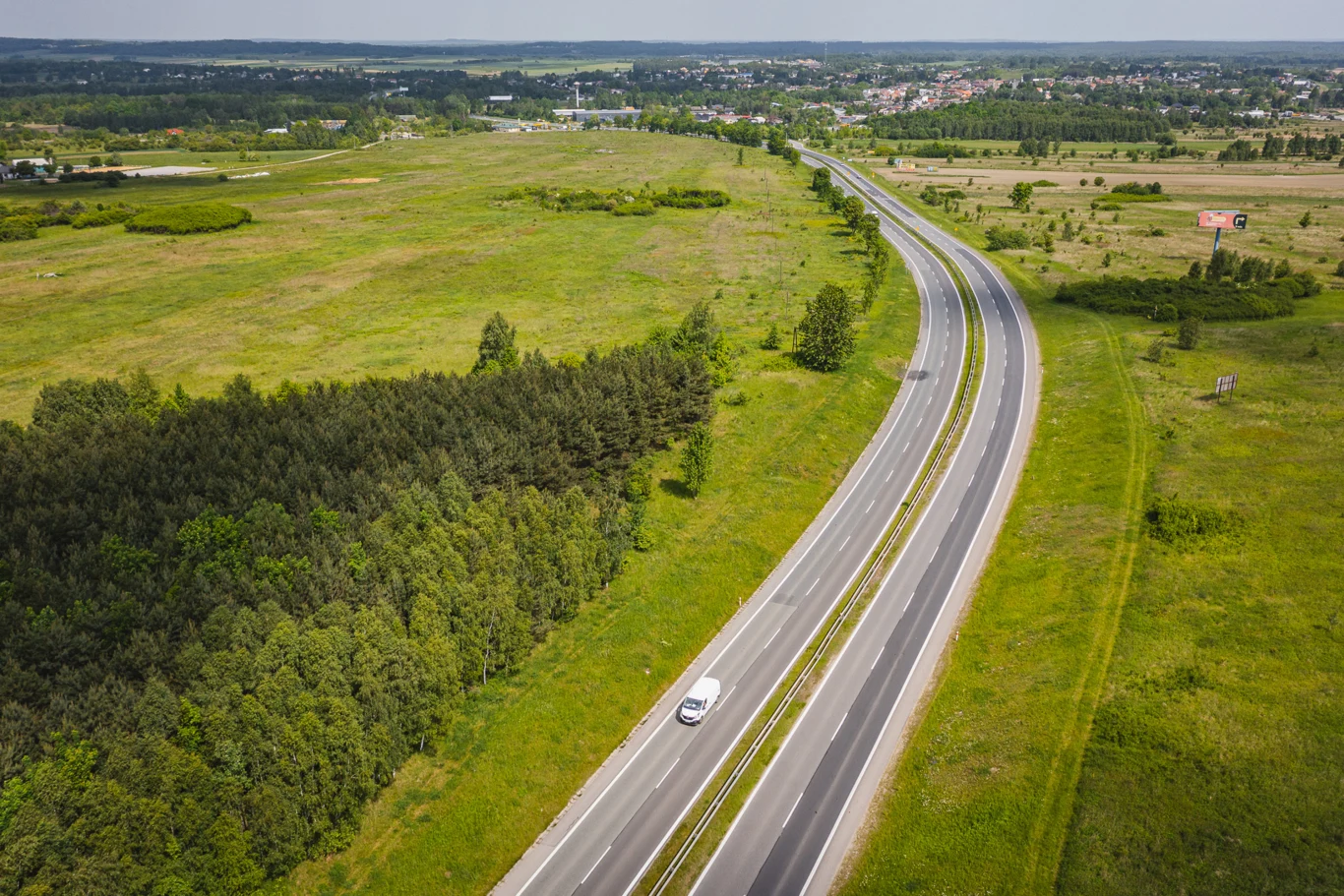Ważny fragment DK91 do przebudowy. GDDKiA podpisała umowę Fragment DK91 przed przebudową, dwupasmowa droga przez pola i las, biały samochód, zabudowa w tle