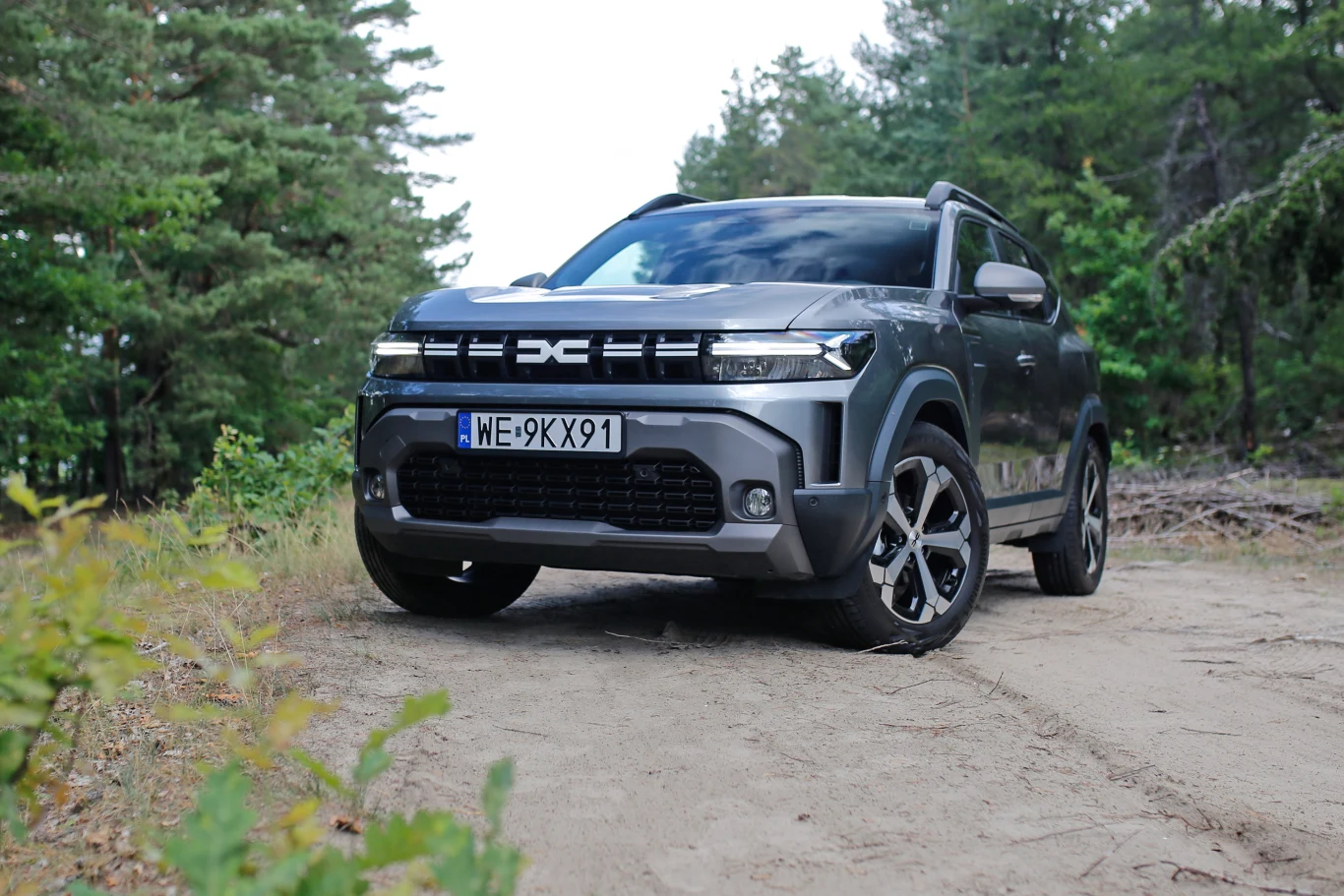 Nowoczesny, szary SUV z czarnymi felgami porusza się po leśnej, piaszczystej drodze, otoczony drzewami iglastymi.