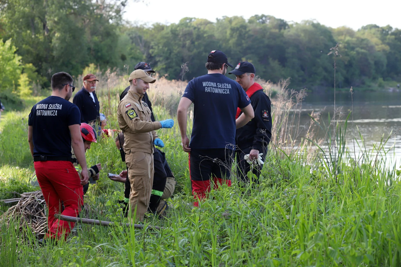 Grupa ratowników wodnych i służb mundurowych prowadzi akcję poszukiwawczą nad brzegiem rzeki, otoczona wysoką trawą i roślinnością.