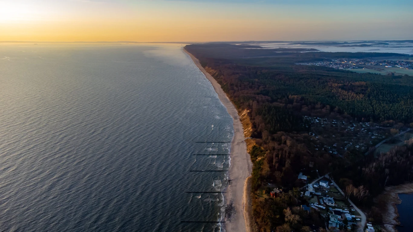Skrawek wyspy Uznam wystawiono na sprzedaż. Linia brzegowa wyspy Uznam z piaszczystą plażą, lasem i widocznymi zabudowaniami, oświetlona zachodzącym słońcem.