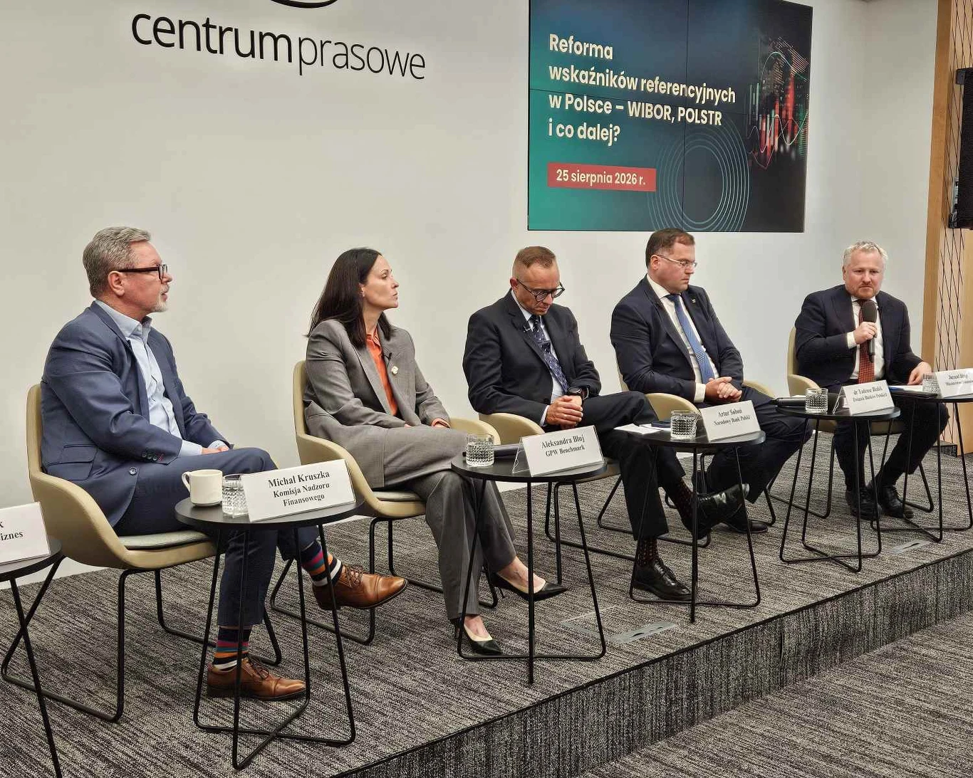 Pięcioro prelegentów siedzi w rzędzie na scenie podczas panelu dyskusyjnego o reformie wskaźników referencyjnych w Polsce, wszyscy ubrani w formalne stroje biznesowe, z mikrofonami i tabliczkami z nazwiskami przed sobą, w tle widoczny baner tematyczny ...