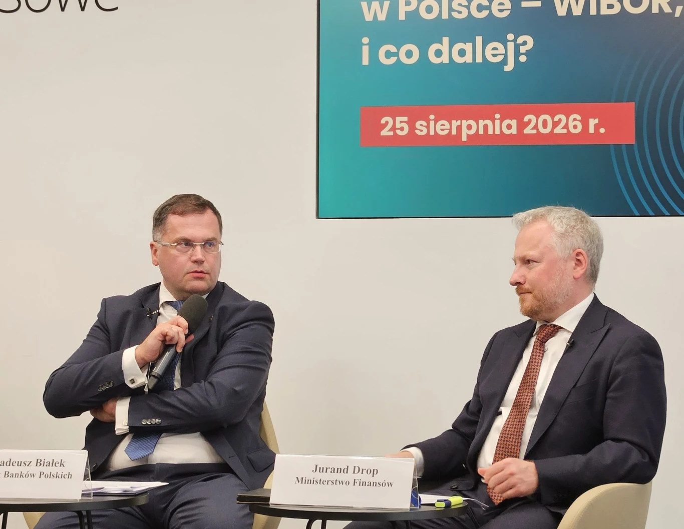 Dwóch mężczyzn w garniturach siedzi przy stole i uczestniczy w dyskusji panelowej, każdy z nich ma przed sobą wizytówkę z imieniem i funkcją, w tle widoczny fragment plakatu informującego o temacie i dacie wydarzenia.