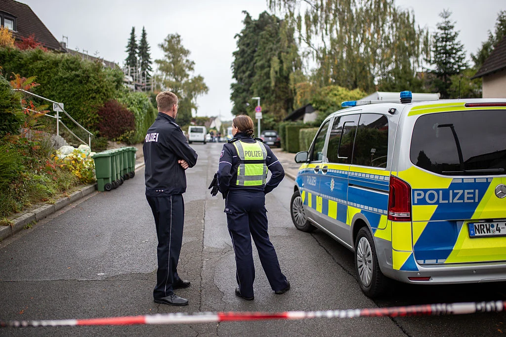 Atak w szkole w Niemczech. Dwie osoby nie żyją (zdj. ilustracyjne) Dwóch policjantów stoi obok radiowozu z włączonymi światłami, ulica odgrodzona taśmą policyjną.