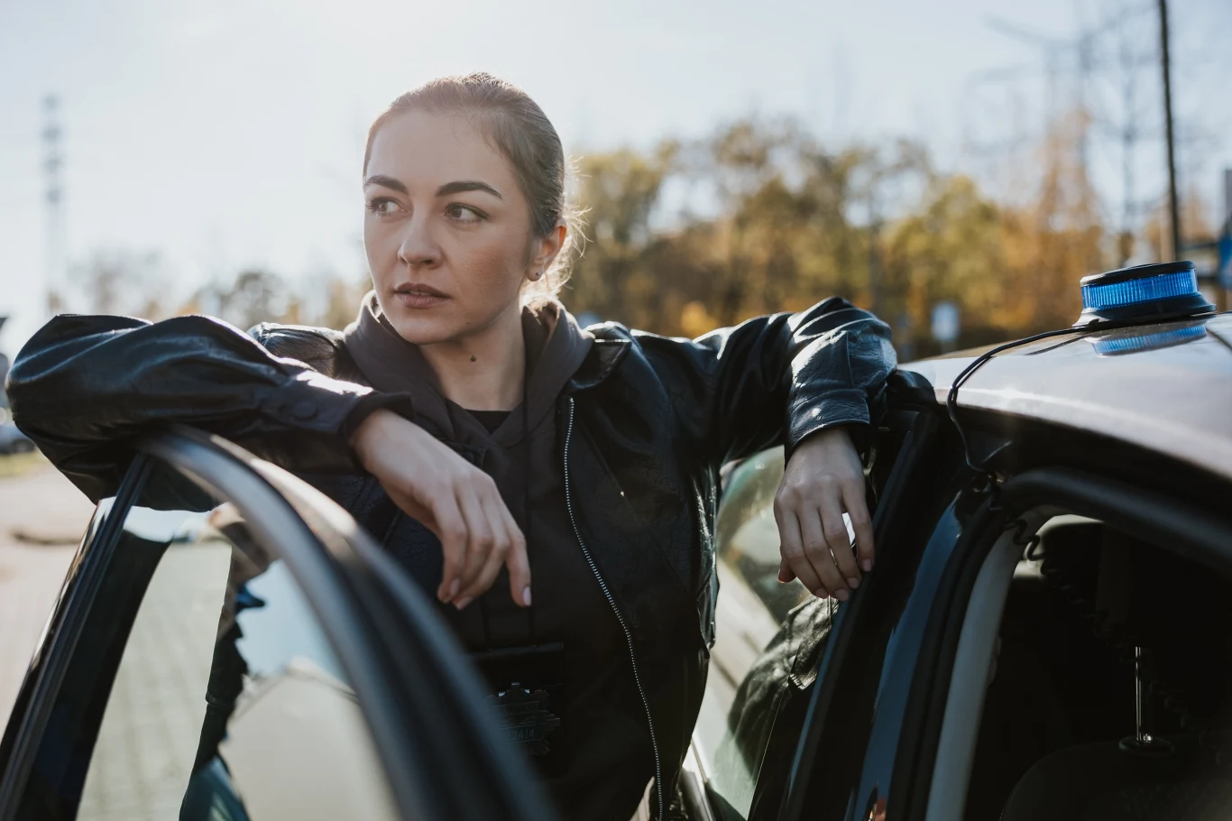 Adrianna Izydorczyk Kobieta w skórzanej kurtce oparta o otwarte drzwi radiowozu policyjnego, z poważnym wyrazem twarzy i skupionym spojrzeniem skierowanym w bok.