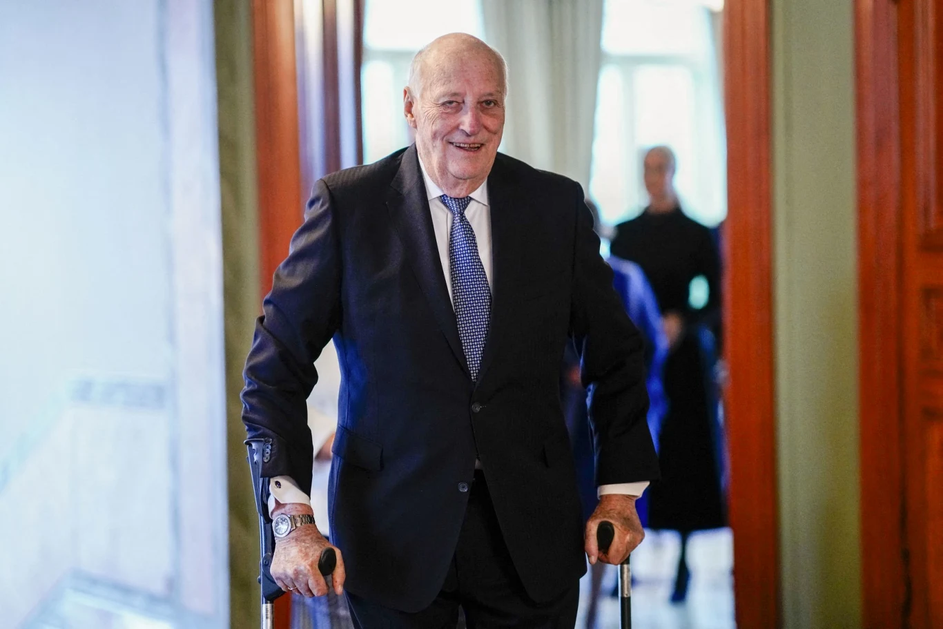 Król Norwegii Harlad V znajduje się w bardzo ciężkim stanie w szpitalu Król Norwegii Harald V w garniturze i krawacie stoi z kulami ortopedycznymi w eleganckim wnętrzu.