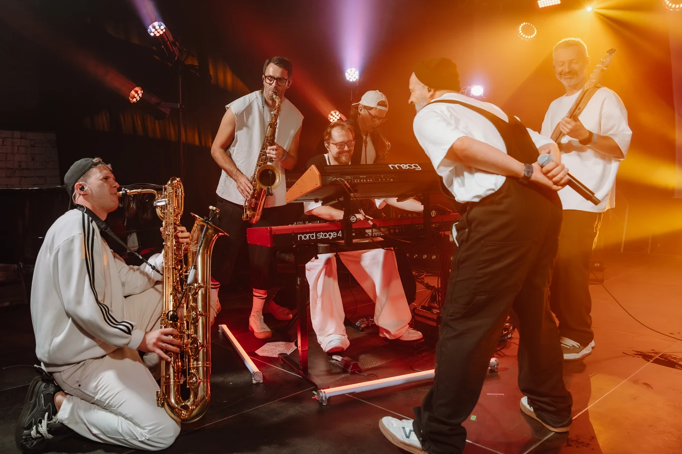 Bibobit Bibobit – pięciu muzyków na scenie, saksofoniści, klawiszowiec, gitarzysta i wokalista w jasnych ubraniach, kolorowe światła.