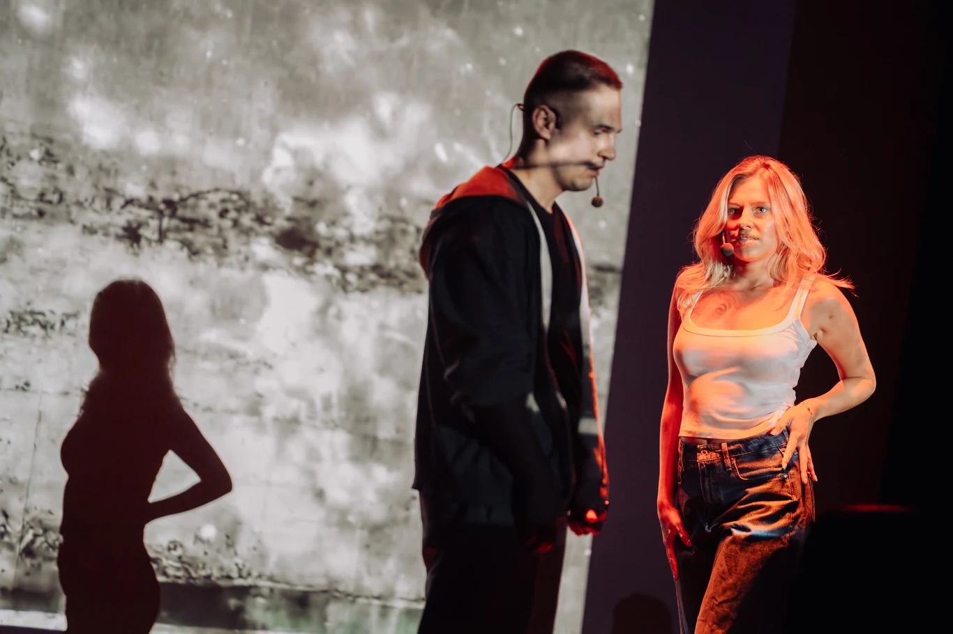 Spektakl "Chłopcy" Dwoje młodych ludzi na scenie teatralnej, z wyraźnie widocznymi emocjami na twarzach, w kontrastowym oświetleniu; kobieta w białym topie i spodniach stoi z boku, mężczyzna w ciemnej bluzie odwraca głowę, pomiędzy nimi napięcie i wyraźna różnica nastrojów.