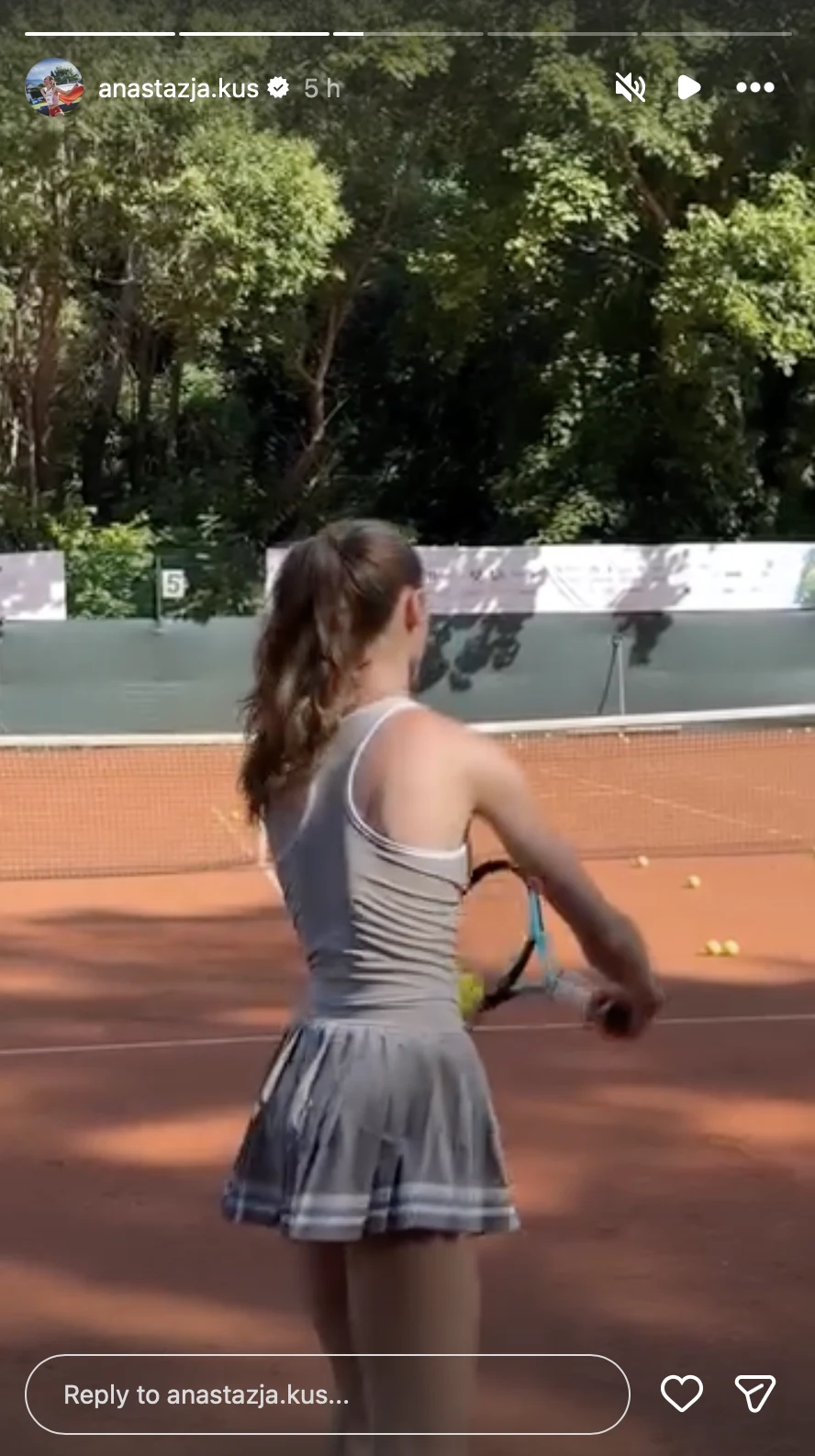 Młoda dziewczyna w sportowym stroju stoi na korcie tenisowym z rakietą w dłoni, przygotowując się do zagrania uderzenia; na ziemi leżą rozrzucone piłki tenisowe, a w tle widoczna jest zielona roślinność.