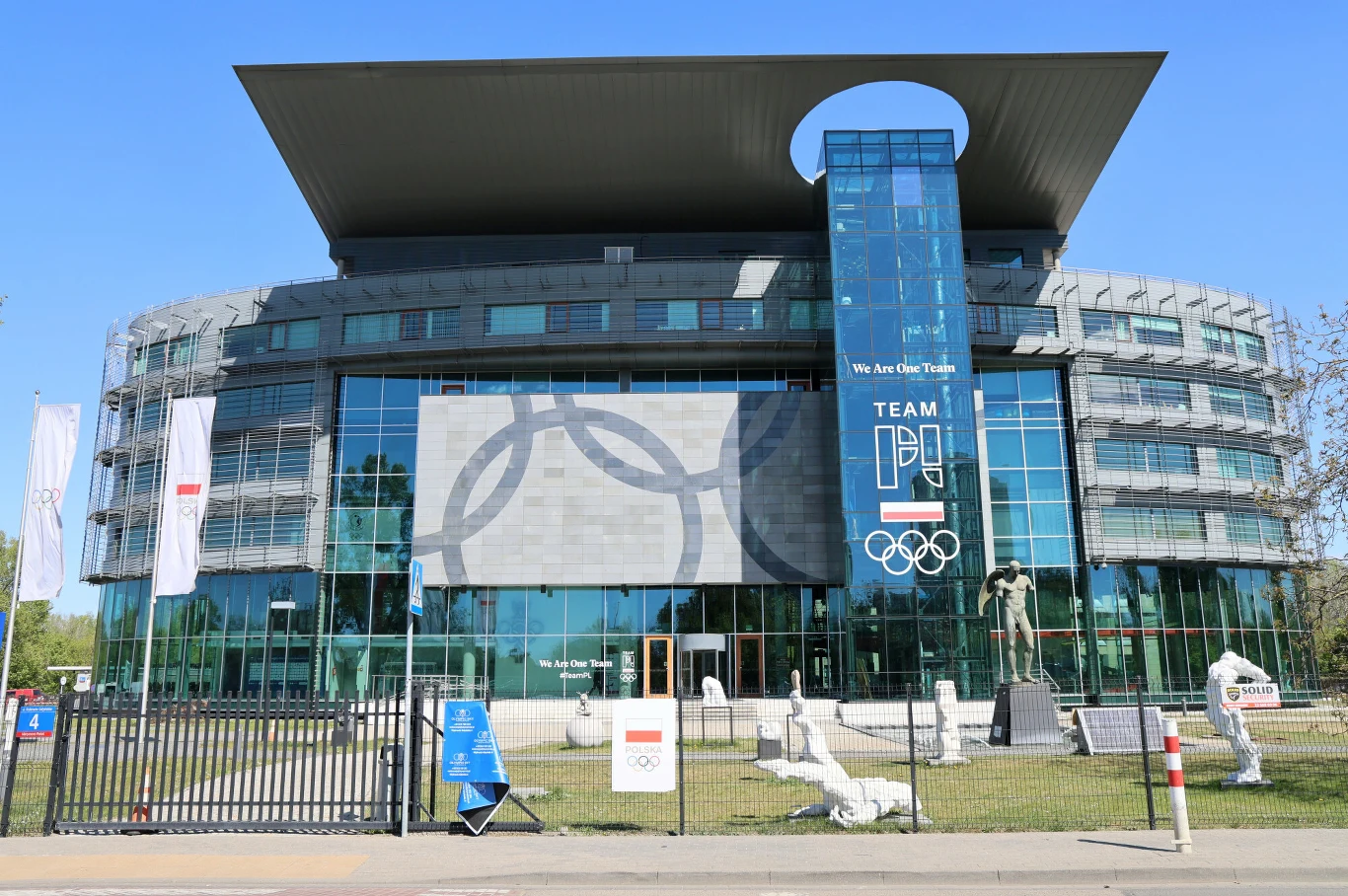 Media: Prezes PKOI usłyszał zarzuty w związku z aferą Zondacrypto Szklany budynek z dużym, prostokątnym dachem, banerem z olimpijskimi symbolami i napisem TEAM POLSKA oraz rzeźbami sportowców przed wejściem.