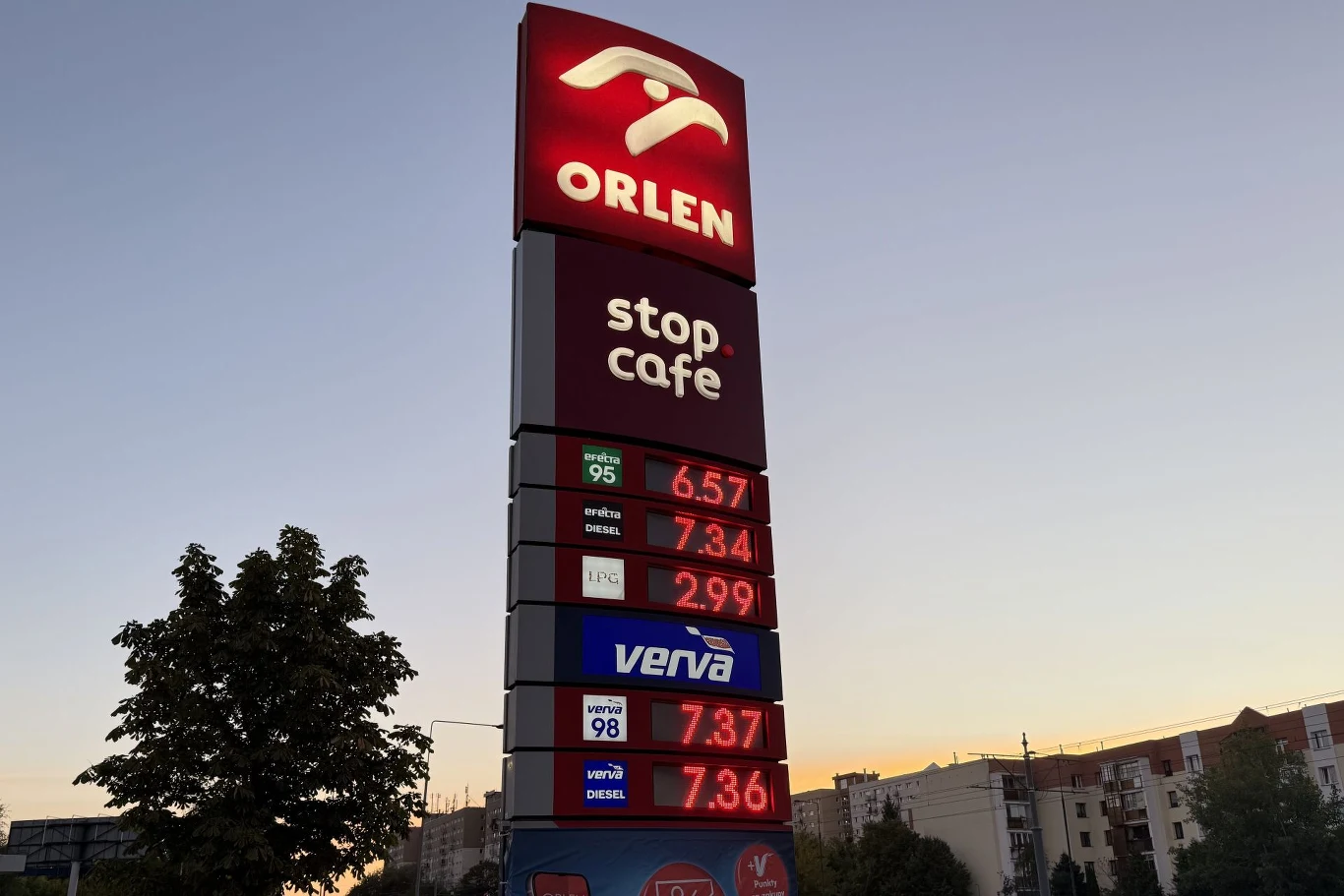 pylon stacji paliw Orlen z wyraźnie widocznymi aktualnymi cenami benzyny, oleju napędowego i LPG na tle wieczornego nieba oraz fragmenty zabudowy miejskiej po prawej stronie