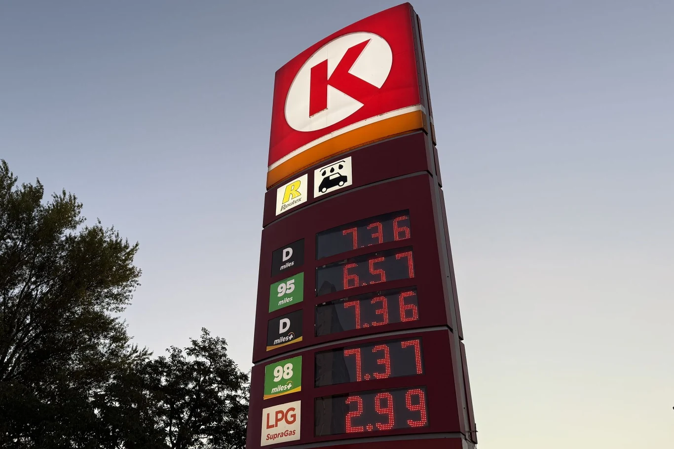 wysoki pylon stacji benzynowej Circle K z wyraźnie widocznymi aktualnymi cenami paliw: ON, benzyny 95, benzyny 98 i LPG, na tle jasnego wieczornego nieba oraz częściowo widocznymi koronami drzew