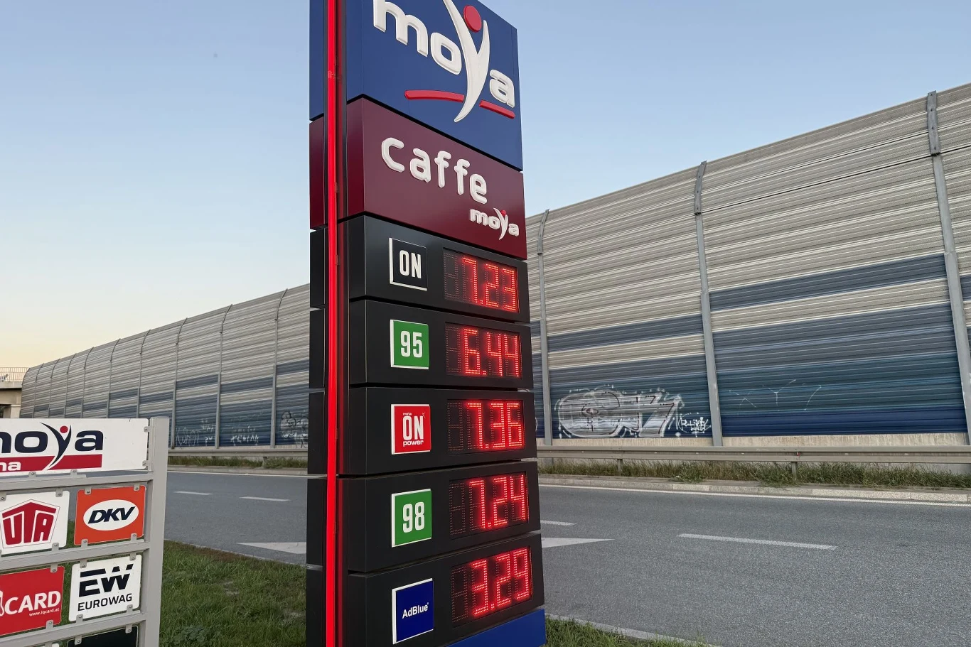 Tablica informacyjna stacji paliw Moya z wyraźnie widocznymi cenami różnych rodzajów paliw, umieszczona przy ulicy z ekranem akustycznym w tle.