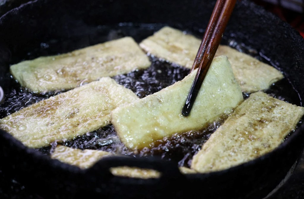 Prostokątne kawałki tofu smażone w głębokim oleju, jeden kawałek podnoszony pałeczkami.