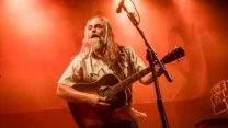Mężczyzna z długimi włosami i brodą grający na gitarze akustycznej na oświetlonej scenie, z mikrofonem skierowanym w jego stronę.