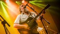 Mężczyzna z długimi włosami i brodą gra na gitarze akustycznej i śpiewa do mikrofonu podczas koncertu, otoczony kolorowym oświetleniem scenicznym.