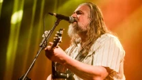 Brodaty mężczyzna z długimi włosami grający na gitarze i śpiewający do mikrofonu na scenie oświetlonej ciepłym światłem.